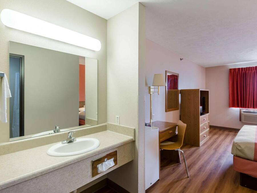 Une chambre d’hôtel avec un grand miroir, un lavabo, un bureau, une chaise, un lit et une fenêtre avec des rideaux.