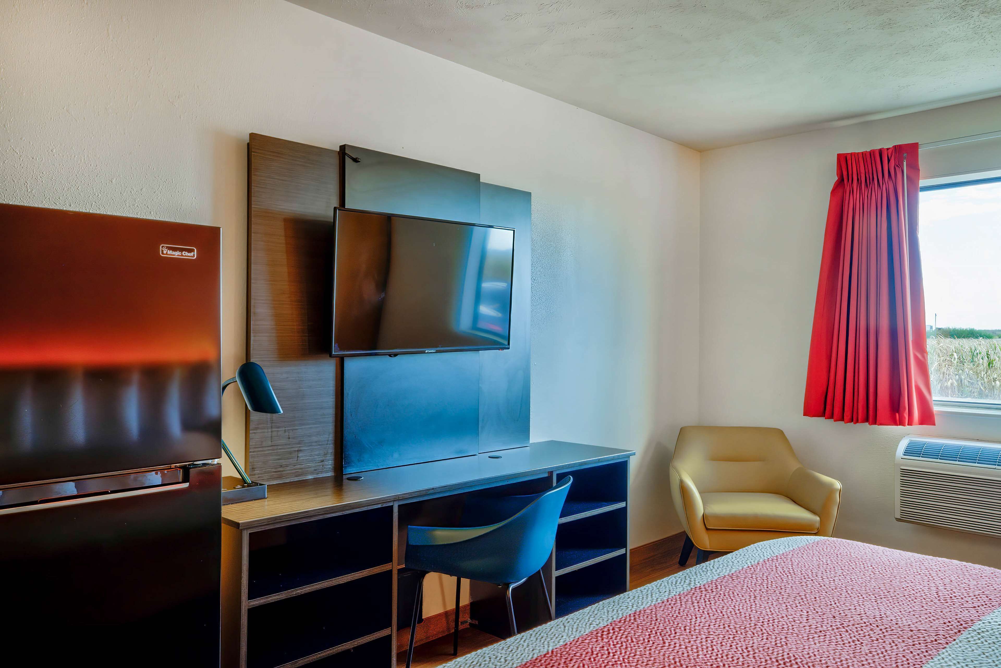 A hotel room with a flat-screen TV mounted on the wall, a desk with a chair, a bed with a red and white striped blanket, and a window with red curtains.