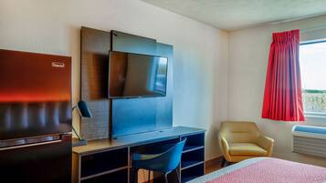 Habitación de hotel con televisión de pantalla plana en la pared, escritorio con silla, cama con manta a rayas rojas y blancas, y ventana con cortinas rojas.