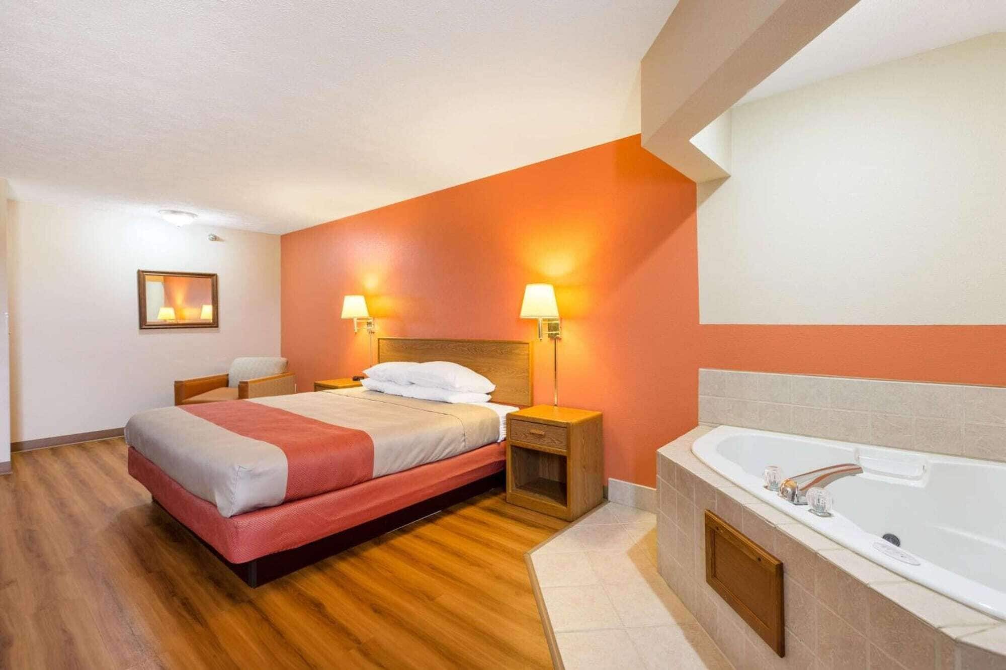A hotel room with a bed, bedside lamps, a nightstand, a wooden headboard, a large window, and a bathtub.