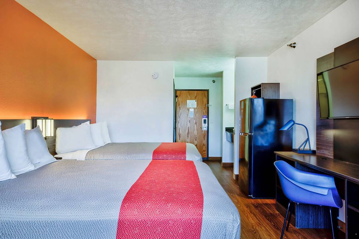 Ein Hotelzimmer mit zwei Betten, einem Schreibtisch, einem Stuhl, einem Kühlschrank und einem Fernseher.