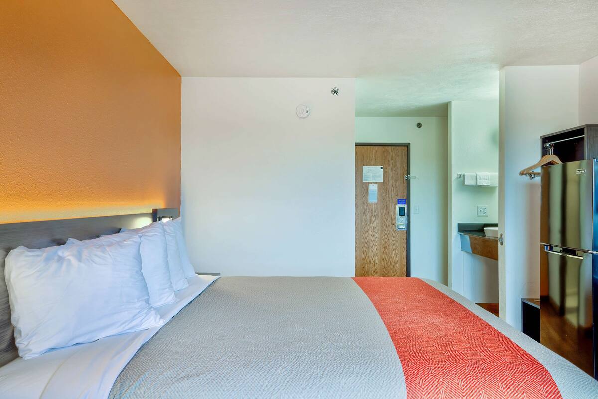 Ein Hotelzimmer mit einem Bett, einem roten und weißen Teppich, einem Holz-Kopfteil, einer weißen Wand, einer Tür zu einem Badezimmer und einem Kühlschrank.