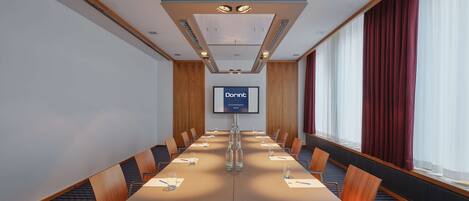 A conference room with a long table, chairs, a projector screen, and a large window with curtains.