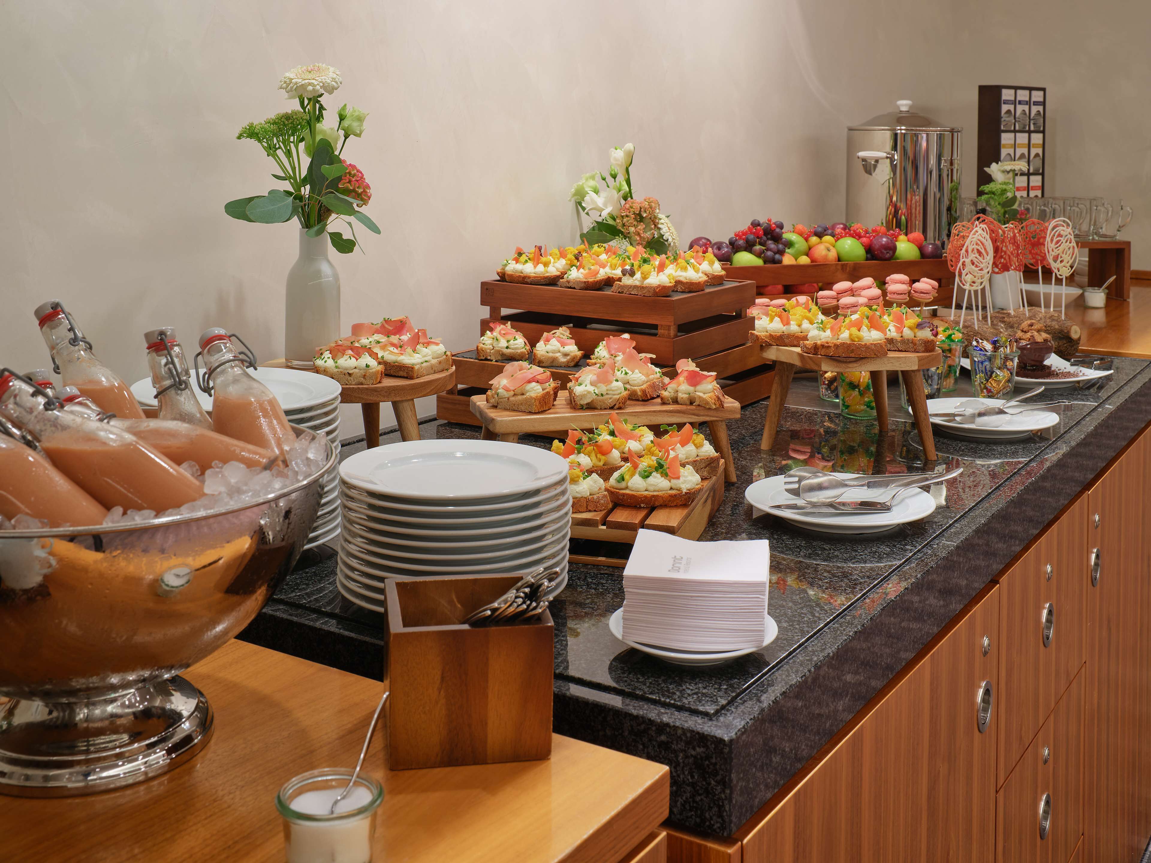 A buffet setup with various food items, including sandwiches, fruits, and desserts, arranged on wooden tables and a marble counter.