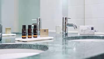 A bathroom with two sinks, a mirror, and a shelf with three bottles and a box.