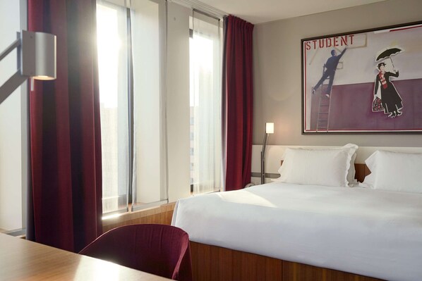 Une chambre d’hôtel avec un lit, un bureau, une chaise, une lampe et une fenêtre avec des rideaux.