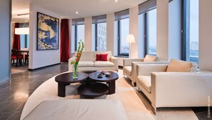 A modern hotel room with a living area featuring a sofa, armchairs, a coffee table, and a dining area with a table and chairs. Large windows provide views of the city. - Dorint Kurfürstendamm Berlin (Berlin)