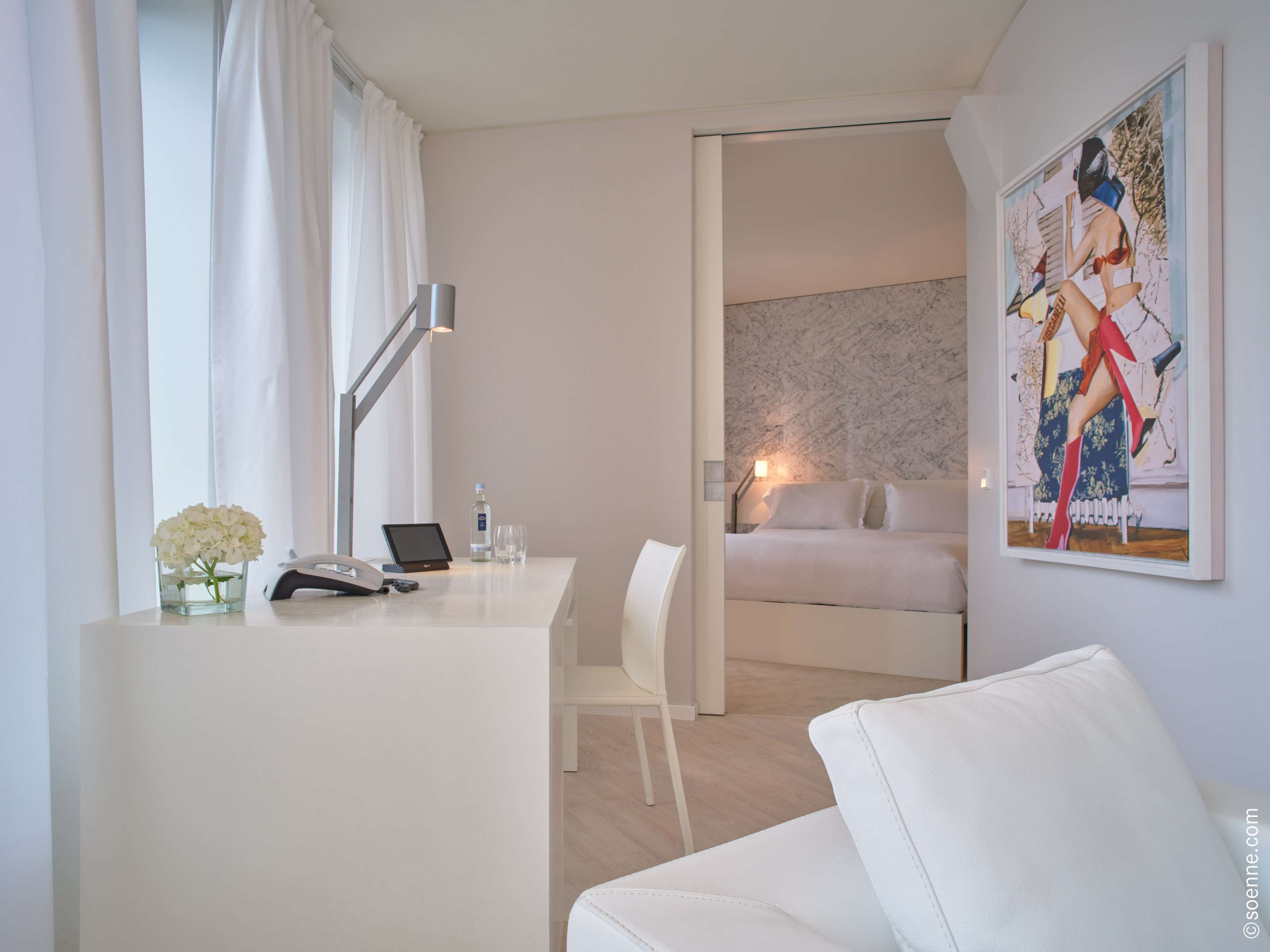 A modern hotel room with a white desk, a laptop, a phone, a vase of flowers, a bed with a white headboard, and a painting on the wall.