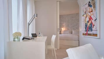 A modern hotel room with a white desk, a laptop, a phone, a vase of flowers, a bed with a white headboard, and a painting on the wall.
