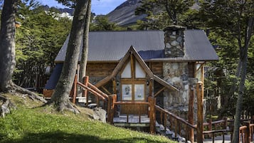 Una cabaña rústica con fachada de piedra, escalera de madera y techo de metal, rodeada de árboles y un paisaje montañoso.