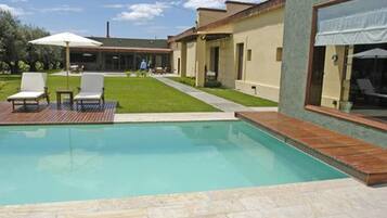 Un área de piscina moderna con una piscina azul cristalina, terraza de madera y dos sillas reclinables blancas.