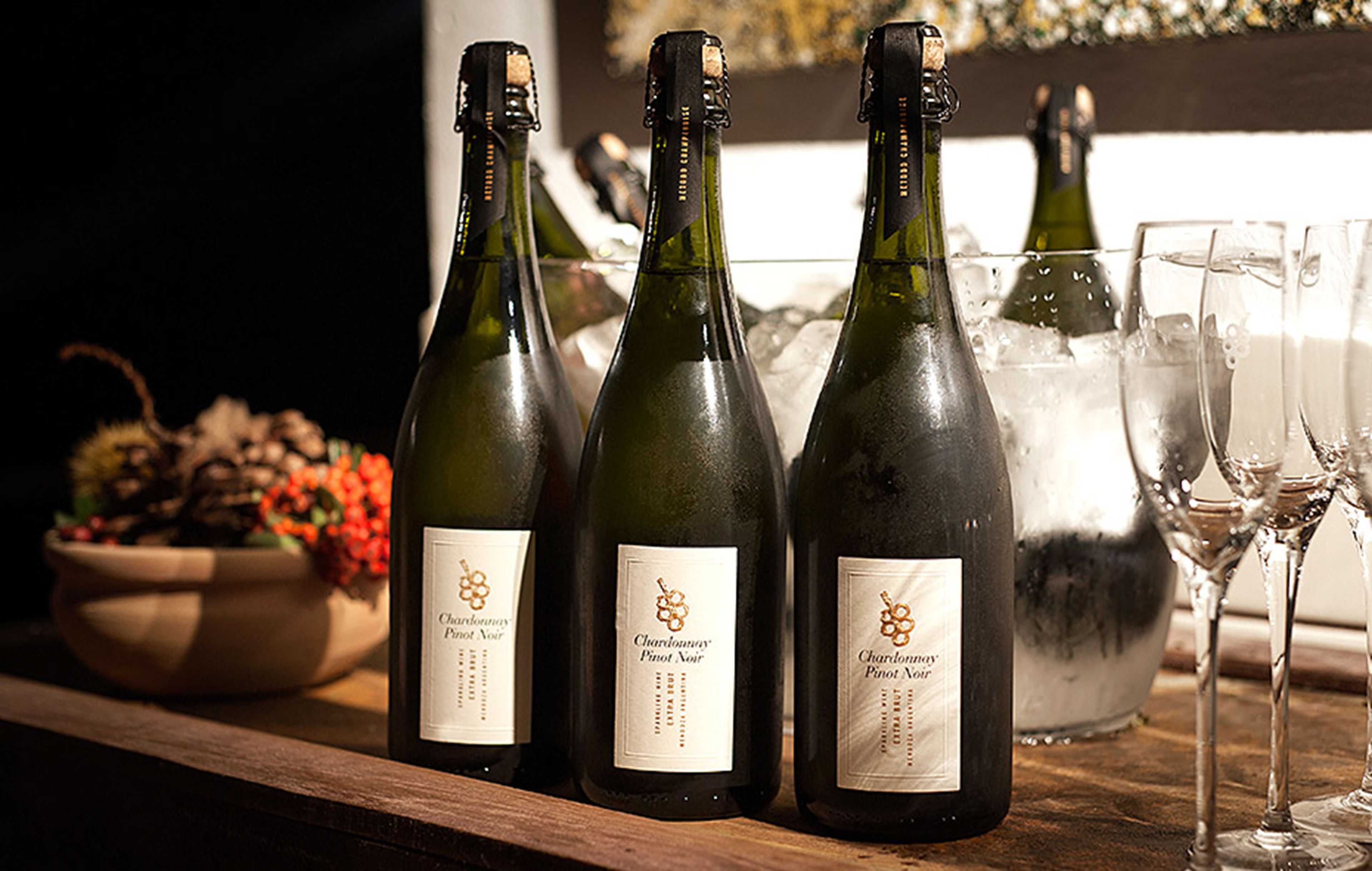Tres botellas de Chardonnay y Pinot Noir sobre una superficie de madera, con copas y un bol de fruta al fondo.