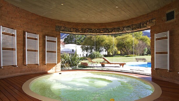 A spa-like room with a hot tub, wooden flooring, stone walls, and a view of a backyard with a pool and greenery. - El Manantial del Silencio (Purmamarca)