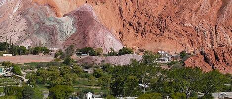 A white building with a flat roof, surrounded by greenery and a large reddish-brown mountain in the background.