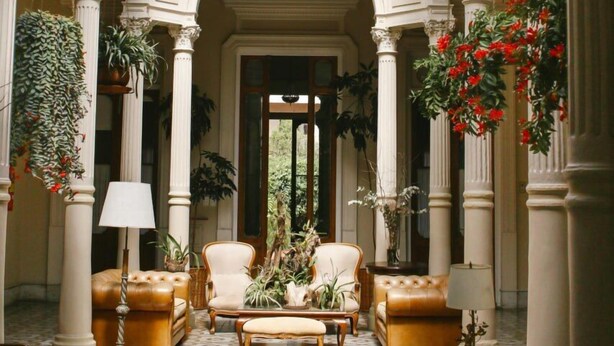 A spacious, elegantly designed interior with a high ceiling and arches, featuring a central seating area with sofas and armchairs, surrounded by potted plants and a patterned floor.