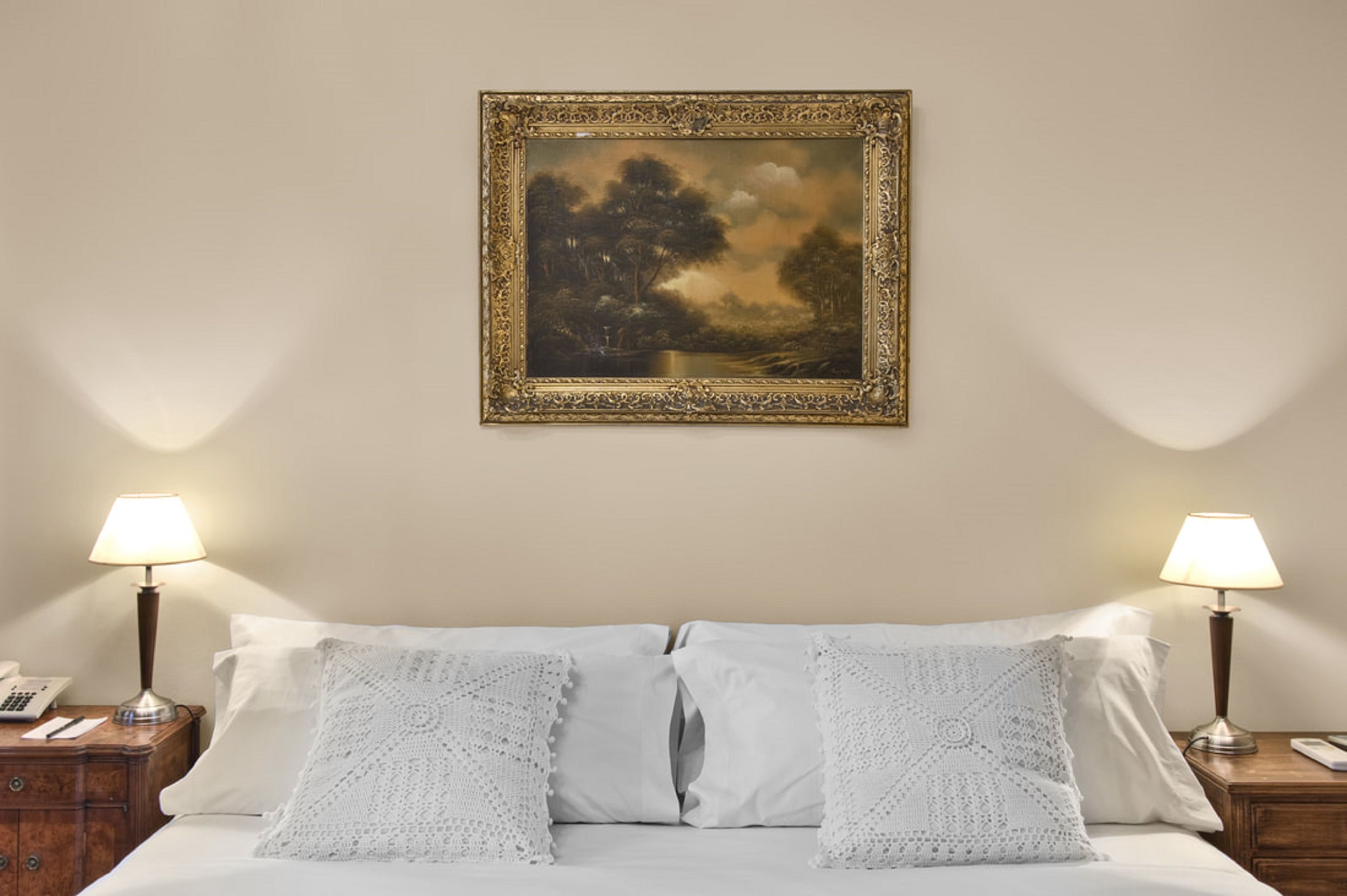 A neatly made bed with white linens and two decorative pillows, flanked by nightstands with lamps and a telephone. A framed painting hangs above the bed.