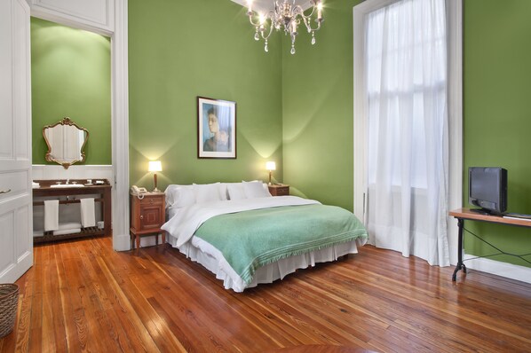 A bedroom with a green wall, wooden floor, a bed with white bedding, a bedside table with a lamp, a mirror, and a television on a stand. - Hotel del Casco (San Isidro)