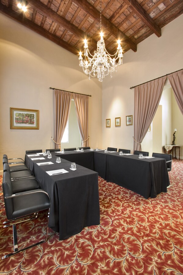Un salón de conferencias con una larga mesa, sillas negras, una araña y una alfombra roja con estampado.