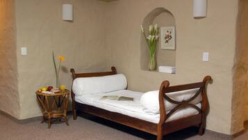 Una cama individual con estructura de madera y ropa de cama blanca, una pequeña mesa redonda con un florero y un espejo con marco en la pared que tiene una imagen floral.