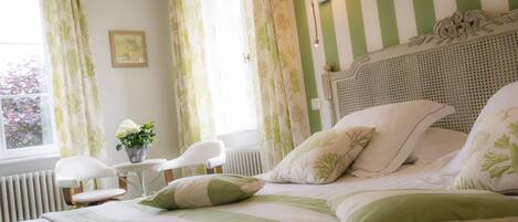 Una cama bien hecha con ropa de cama verde y blanca, una mesita de noche con una lámpara y una silla junto a la ventana con cortinas.
