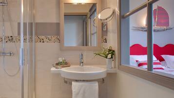 A bathroom with a glass-enclosed shower, a sink with a mirror above it, and a view of a bed with red and white bedding through a window.