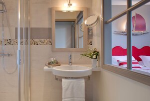 Un baño con ducha de vidrio, un lavamanos con espejo y vistas a una cama con ropa de cama roja y blanca a través de una ventana.