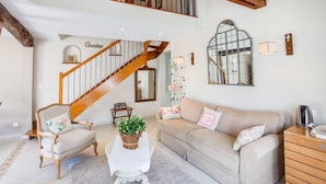 A spacious living room with a high ceiling, a wooden staircase, a beige sofa, a white armchair, and a small table with a plant.