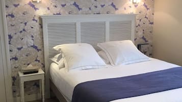 A neatly made bed with white linens and a blue throw, a small bedside table with a remote control, and a wall with floral wallpaper.