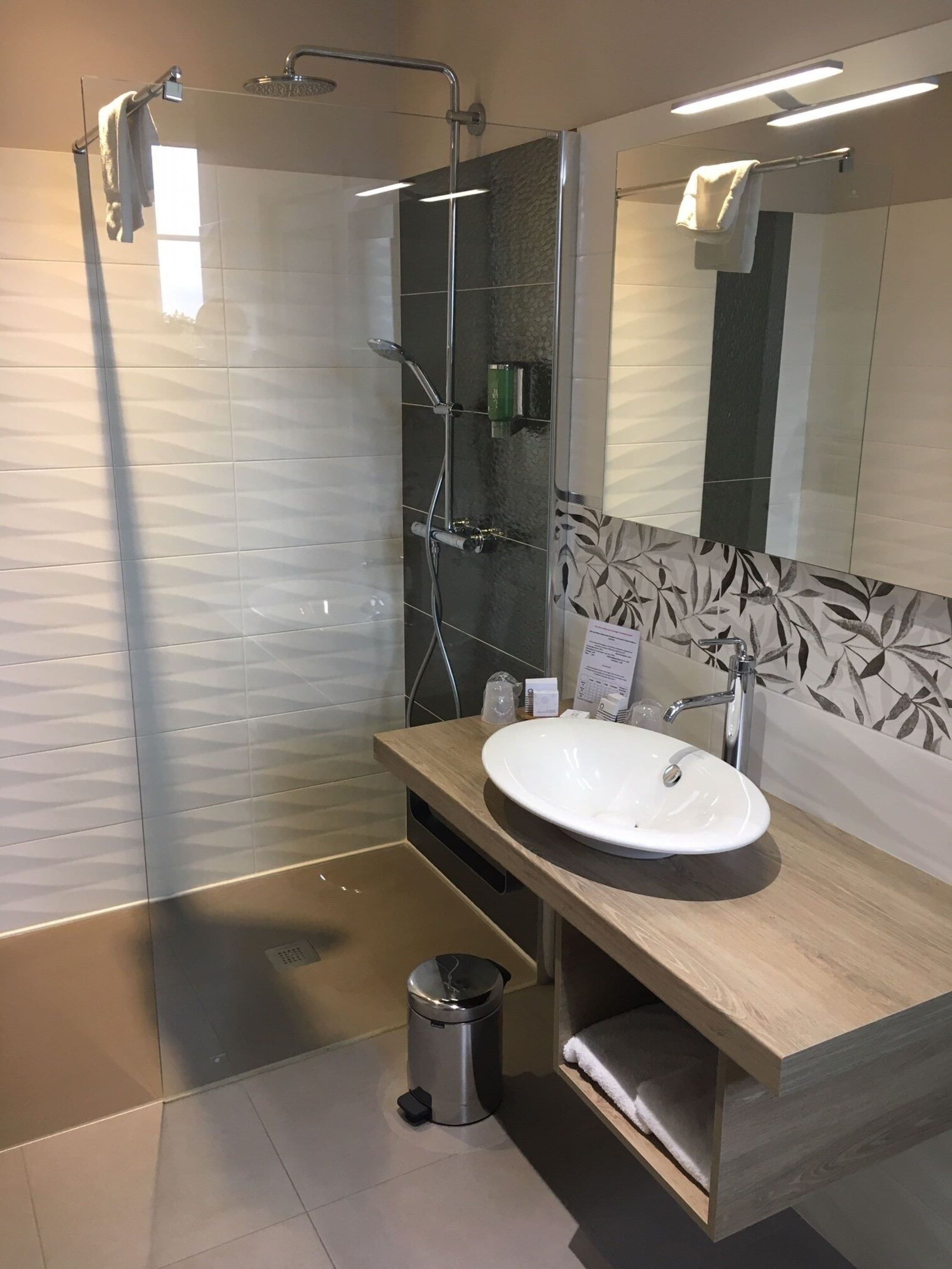 Un baño moderno con ducha de vidrio, tocador de madera y un lavamanos blanco, además de un espejo grande.