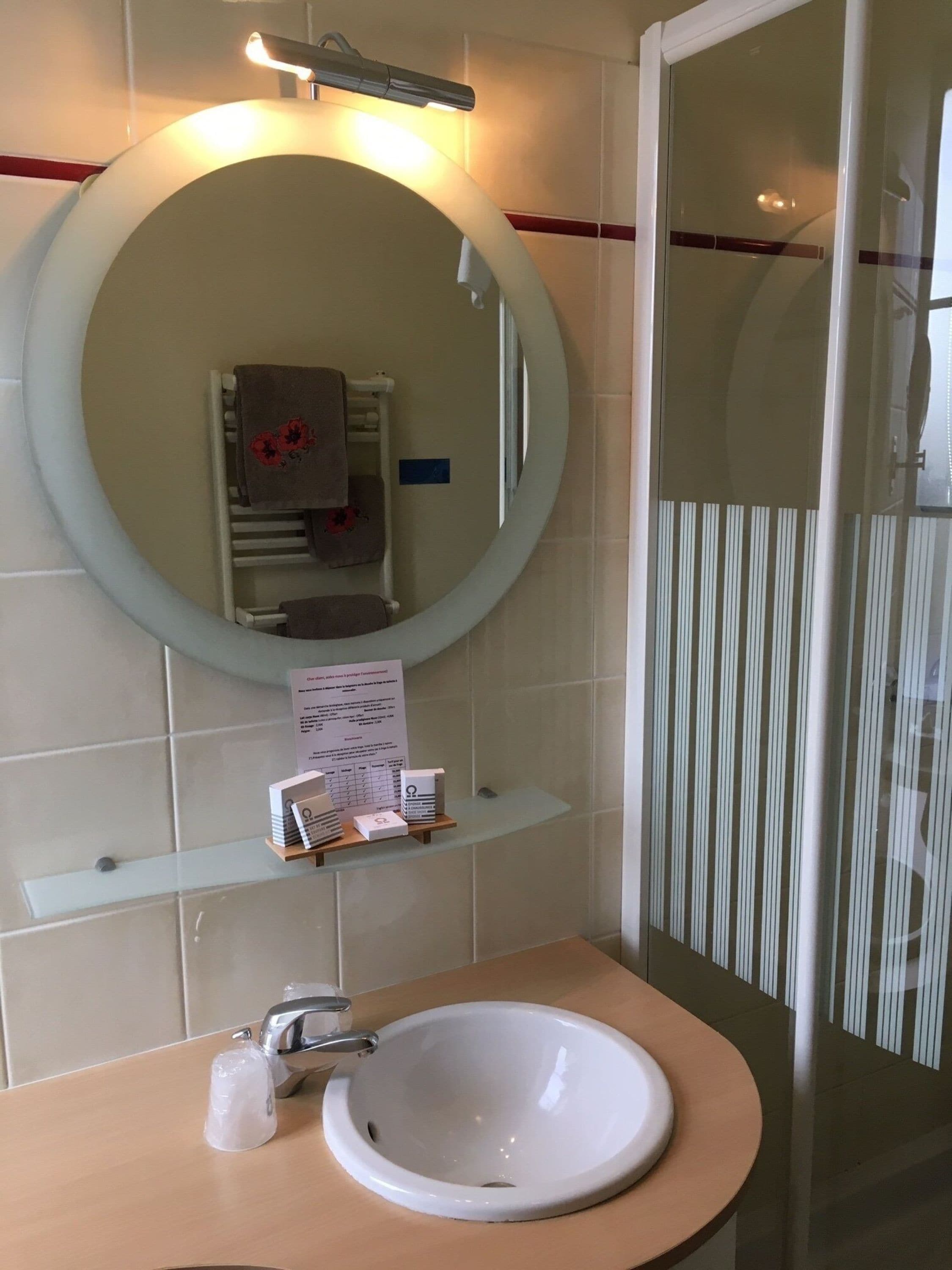 A bathroom with a round mirror, a sink, and a towel rack. There is a sign on the wall and a glass shower enclosure.