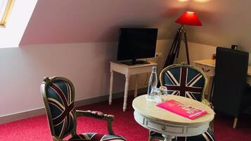 A room with a round table, two chairs with Union Jack designs, a red lamp, a television, and a window with blinds.