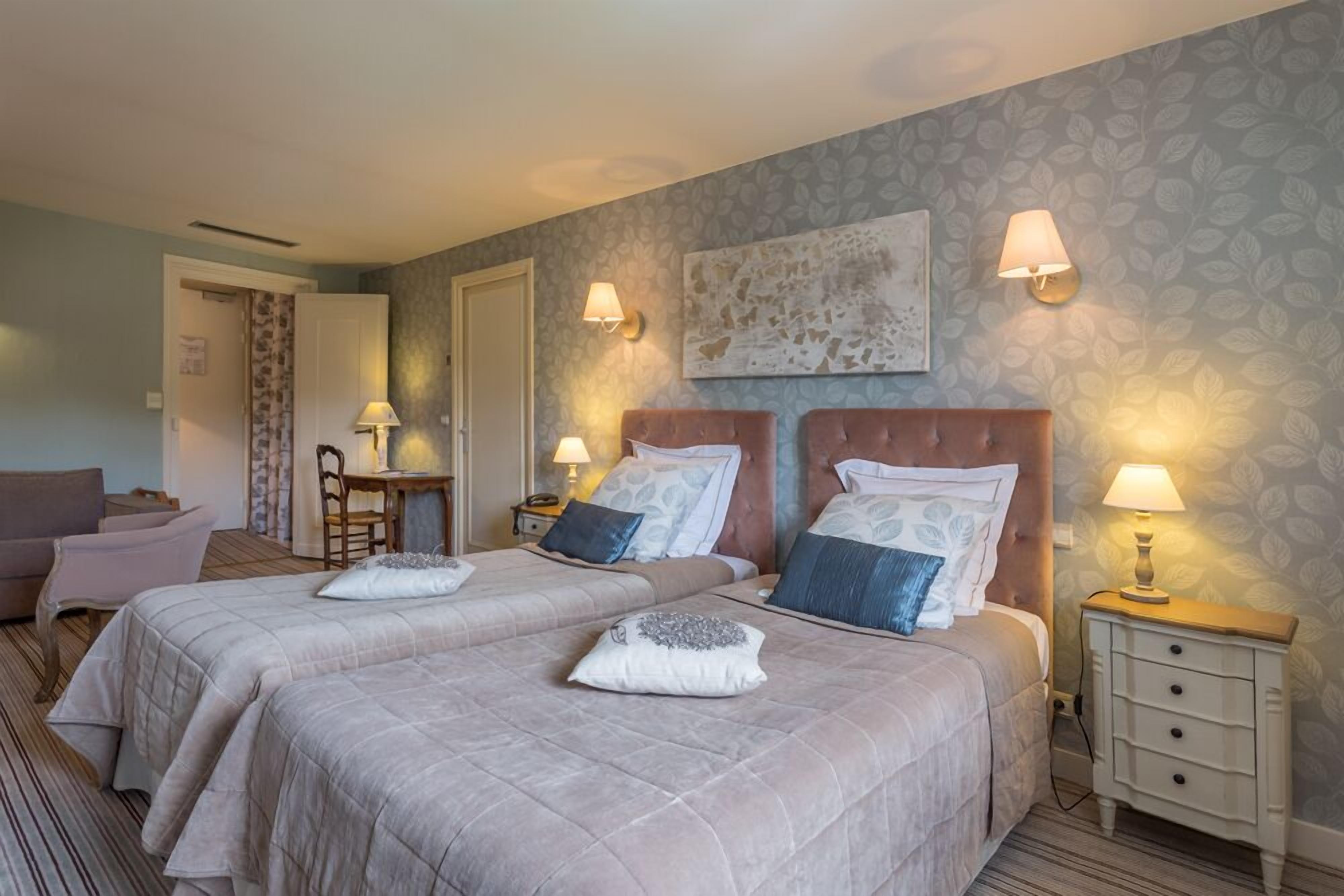Quarto de hotel com duas camas, uma escrivaninha, uma cadeira e uma mesa de cabeceira com luminárias.