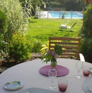 An outdoor dining area with a table set for two, surrounded by greenery and a pool in the background.