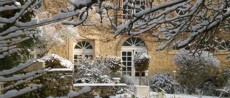 Un patio cubierto de nieve con un edificio de piedra, una fuente y plantas cargadas de nieve.