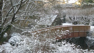 A wooden bridge over a snow-covered garden with trees and a building in the background.