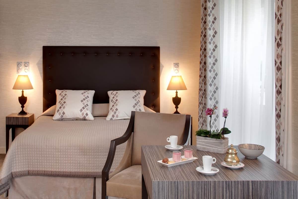 Une chambre d’hôtel avec un grand lit, deux lampes de chevet, une petite table sur laquelle se trouve un plateau de collations et une plante en pot près de la fenêtre.