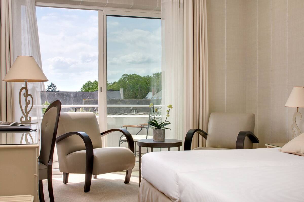 Une chambre d’hôtel avec un grand lit, deux fauteuils, une petite table avec une plante et un bureau avec une lampe.