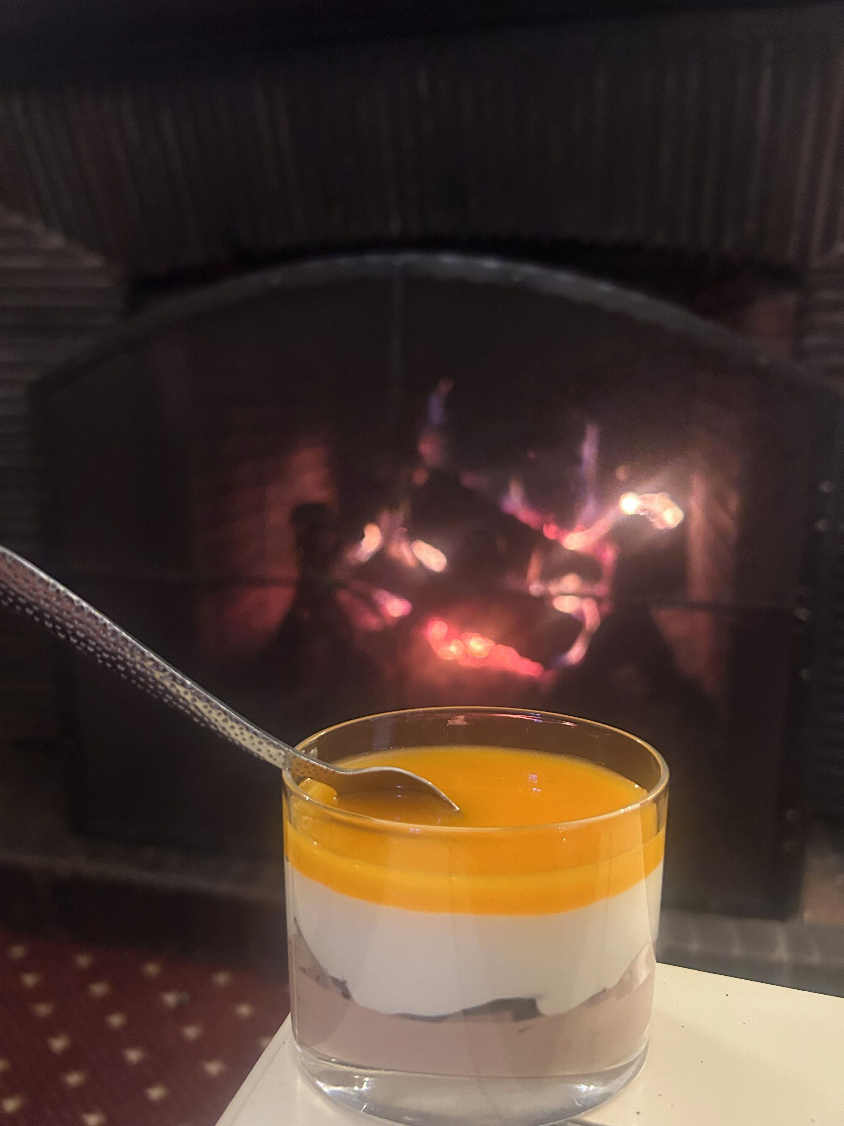 A dessert in a glass with a spoon, set against a background of a fireplace.