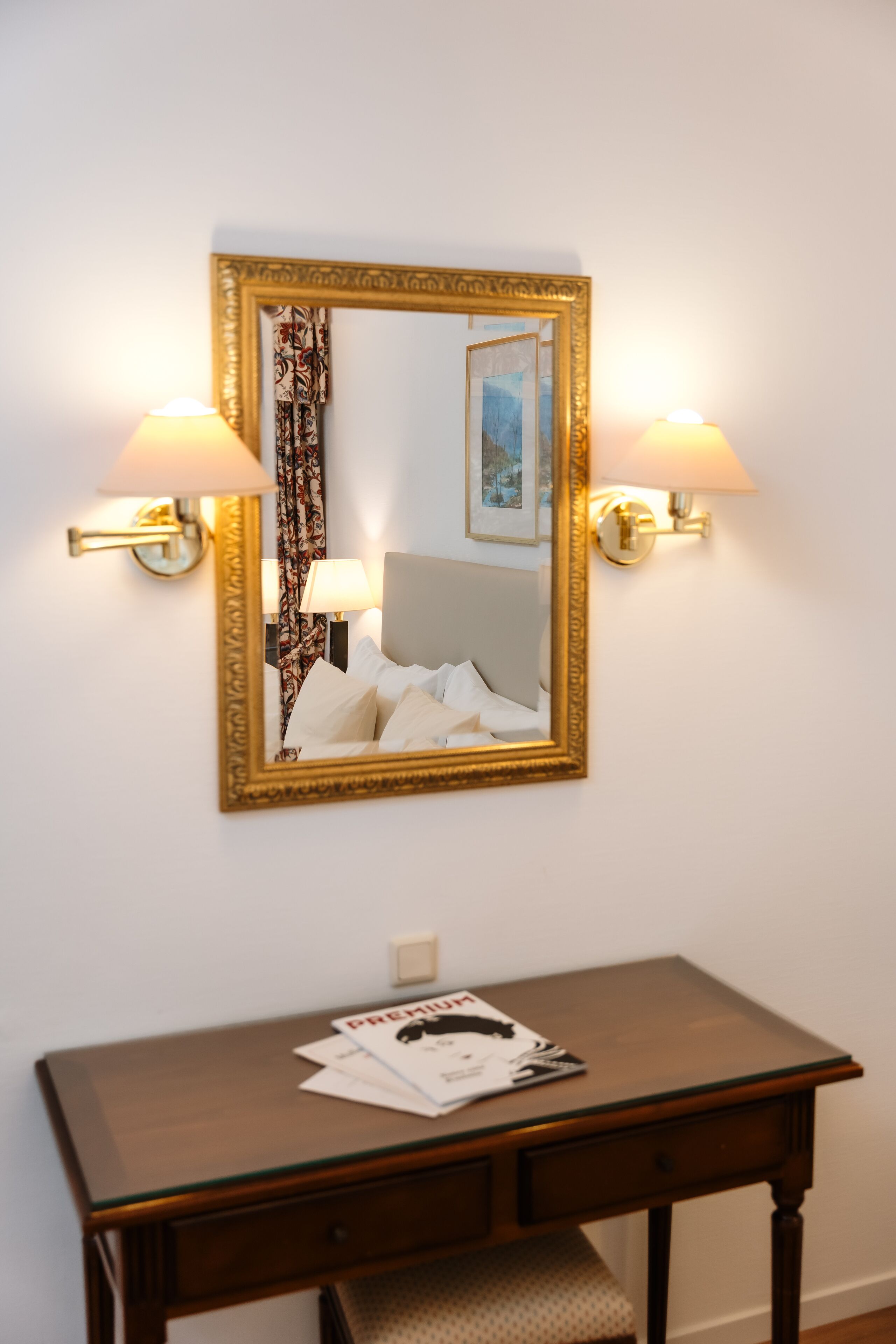Une chambre d’hôtel avec un grand miroir, deux lampes fixées au mur, une table de chevet avec un magazine et un lit recouvert de draps blancs.
