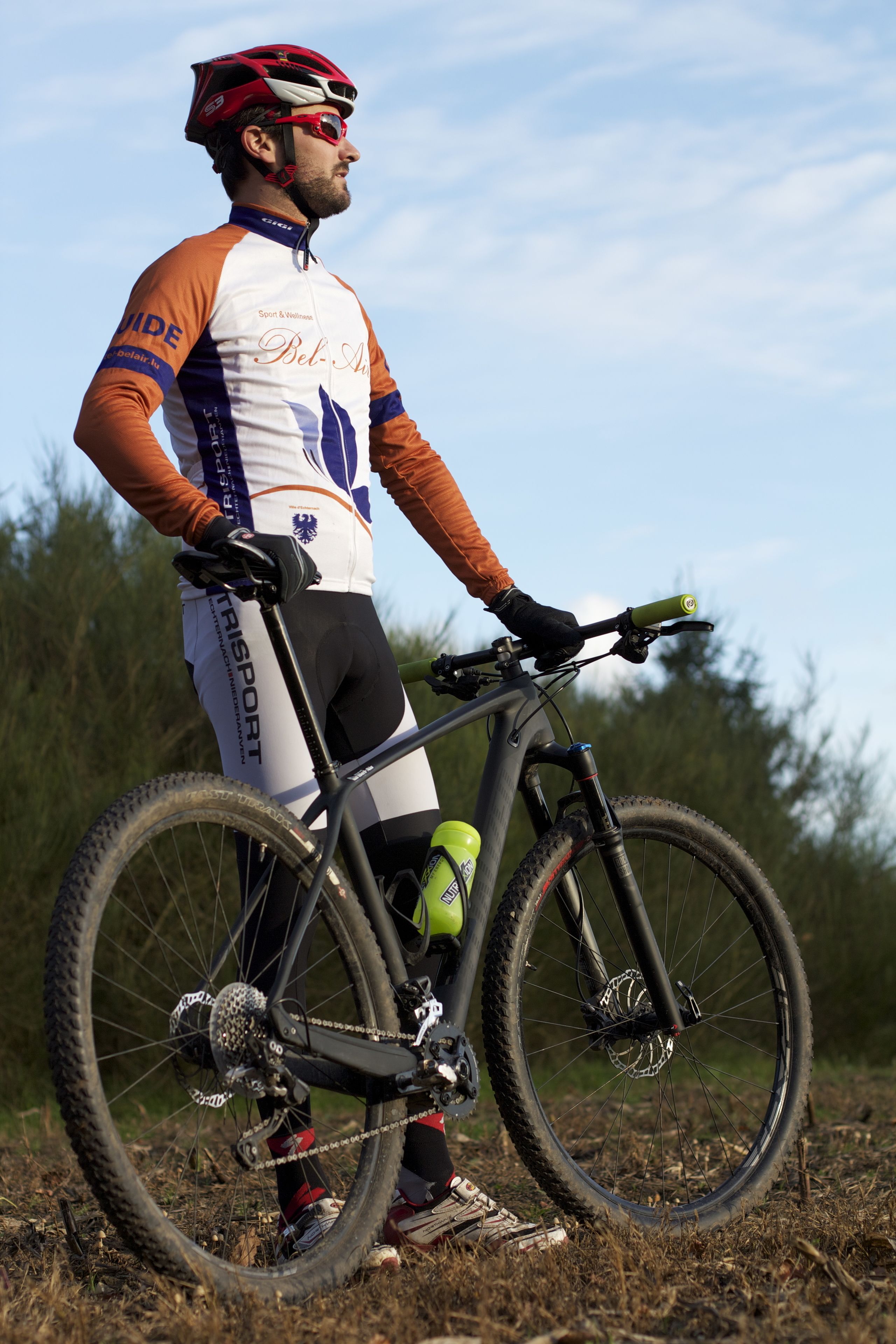 A cyclist in a white and orange jersey with "BEL-AIR" on the chest, standing next to a mountain bike with a green water bottle.
