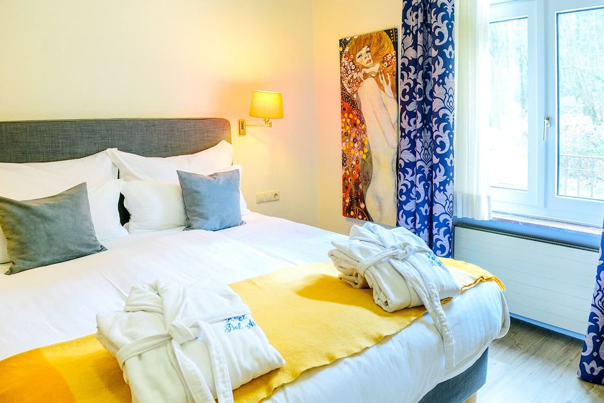 Habitación de hotel con cama, almohadas, lámpara de noche, un cuadro y una ventana con cortinas.