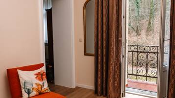 Ein Zimmer mit Holzboden, ein roter Sessel mit einem Blumenkissen, ein kleiner runder Tisch mit Fernbedienung und Zeitschriften, eine Tür mit gemusterten Vorhängen und Blick auf Bäume durch eine Glastür.