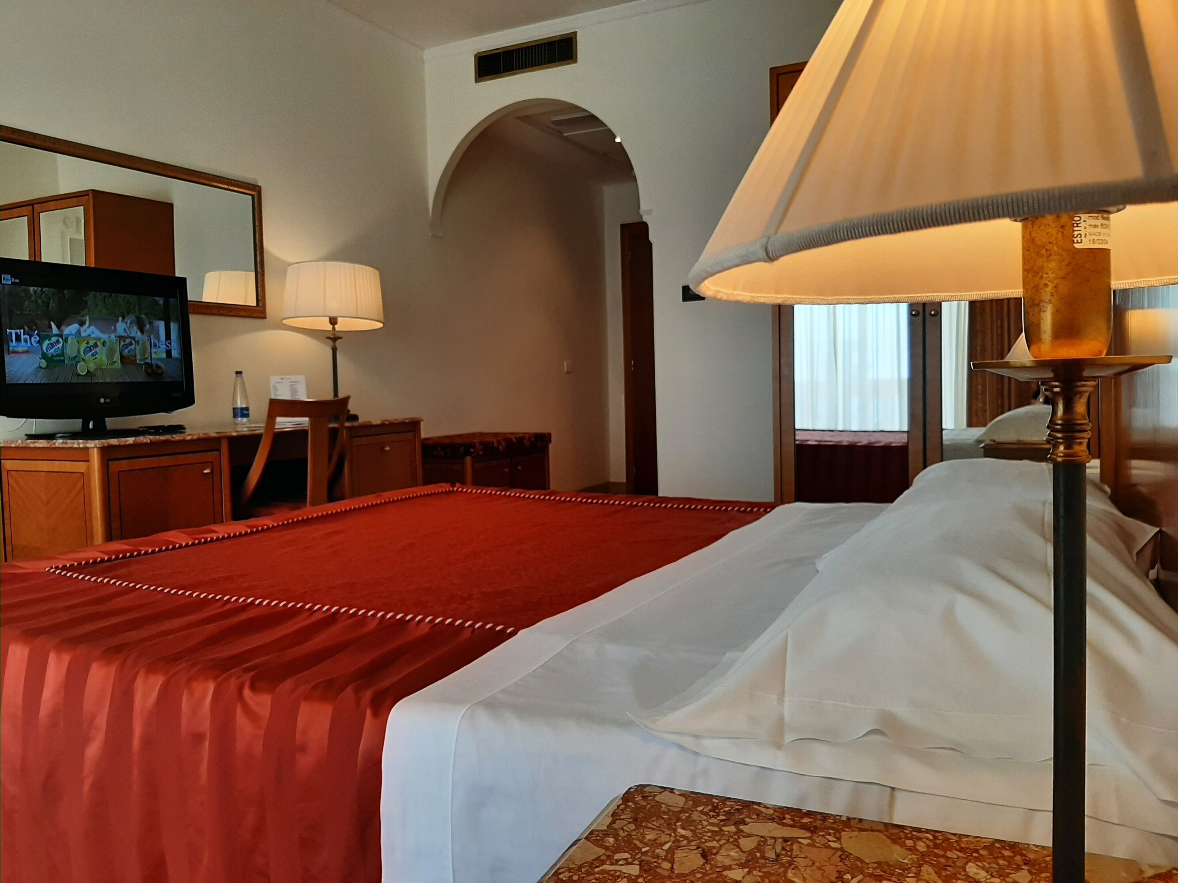 A hotel room with a large bed, a television on a wooden cabinet, a desk with a chair, and a lamp.