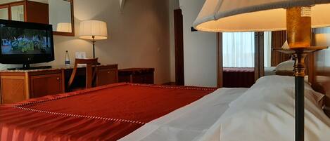 A hotel room with a large bed, a television on a wooden cabinet, a desk with a chair, and a lamp.