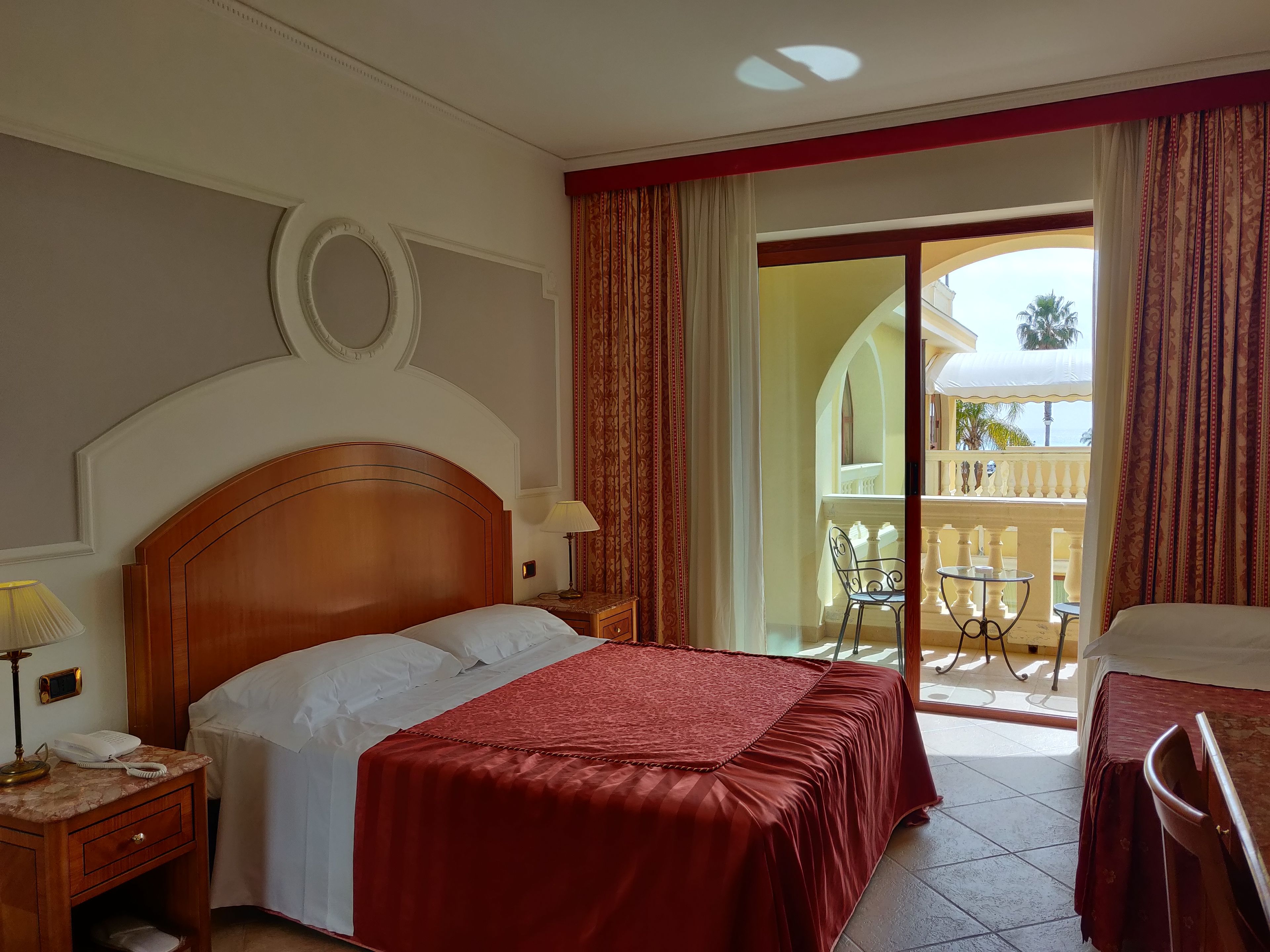 A hotel room with a bed, a desk with a lamp, a chair, and a view of the outside balcony.