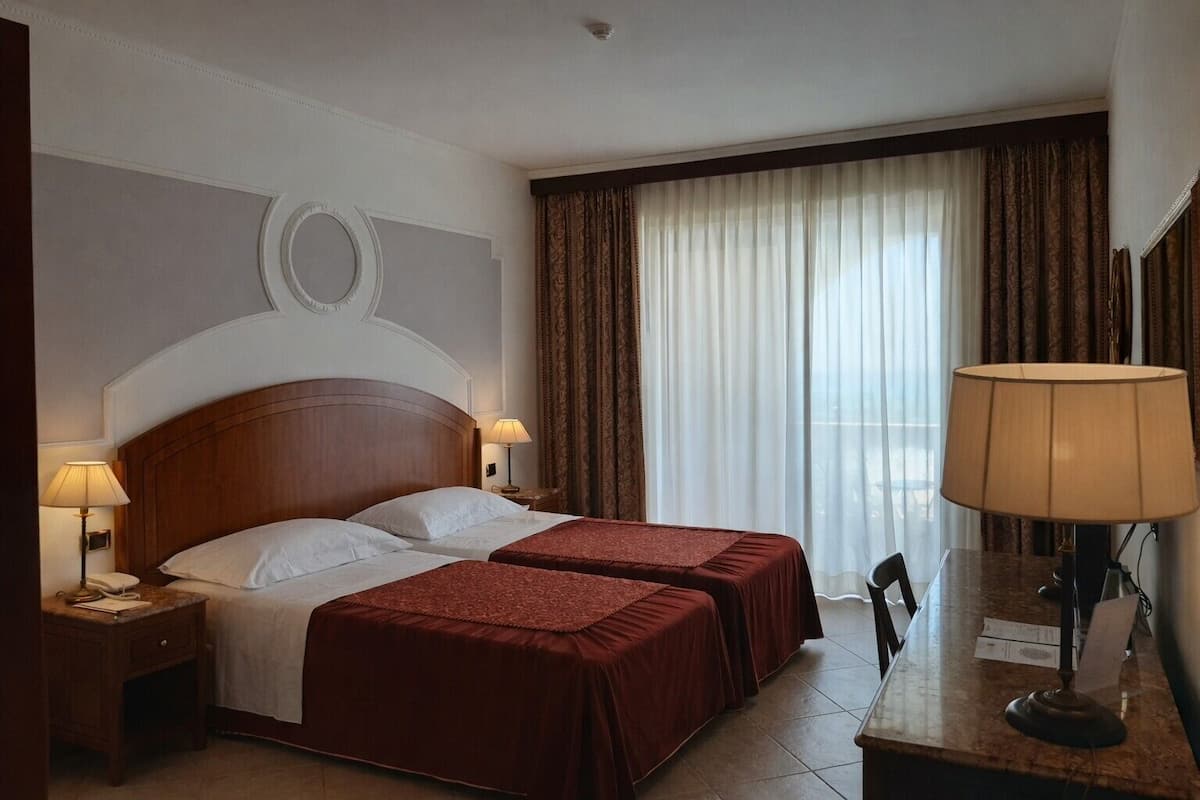 Ein Hotelzimmer mit zwei Betten, einem Schreibtisch, einem Stuhl, einer Lampe und Blick ins Freie durch große Fenster.