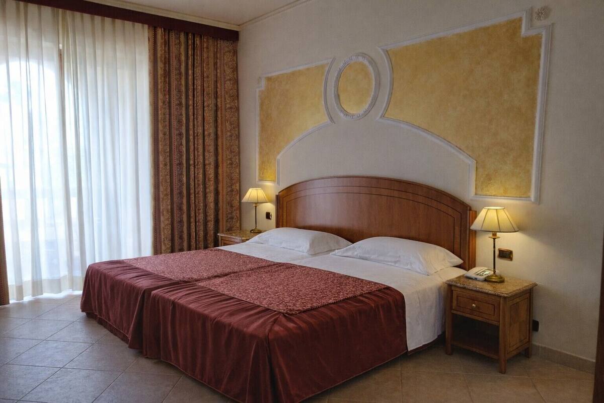 Ein Hotelzimmer mit einem großen Bett, zwei Nachttischen mit Lampen, einem Fenster mit Vorhängen und einem dekorativen Wandbild über dem Bett.