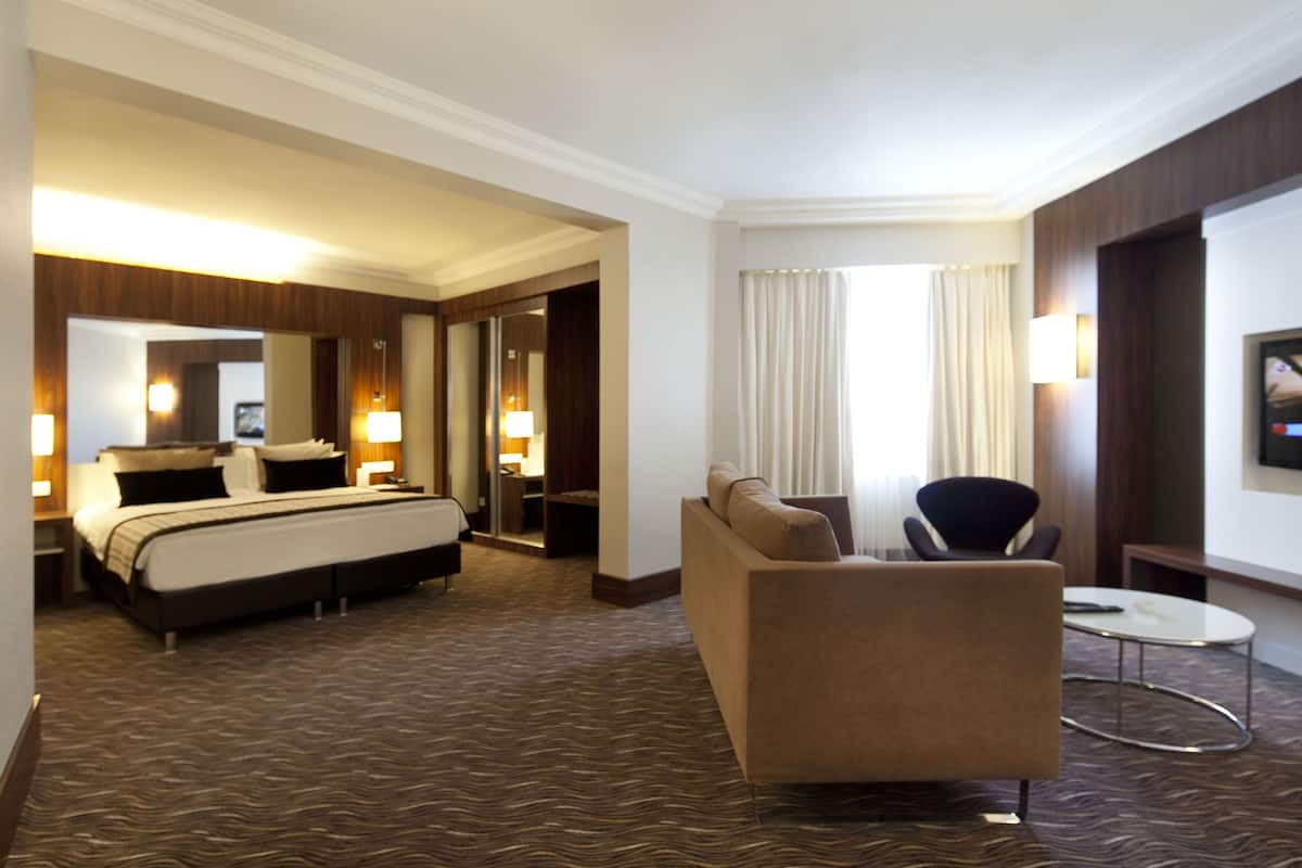 Quarto de hotel com uma cama grande, um sofá, uma mesinha e uma televisão na parede.