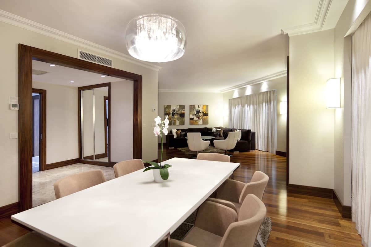 Uma sala de jantar moderna com uma mesa comprida e seis cadeiras, um candelabro, e uma área de estar com um sofá e cadeiras ao fundo.