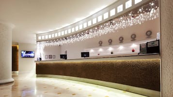 A hotel reception area with a curved reception desk, a wall-mounted clock displaying multiple time zones, and a decorative light fixture above the desk.